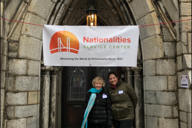 Two female employees smiling & standing in front of a Nationalities Service Center banner