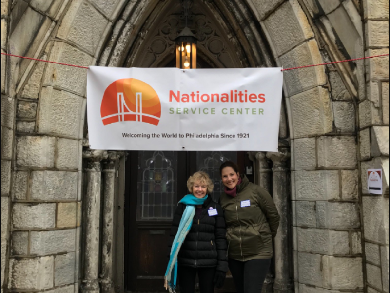 Two female employees smiling & standing in front of a Nationalities Service Center banner